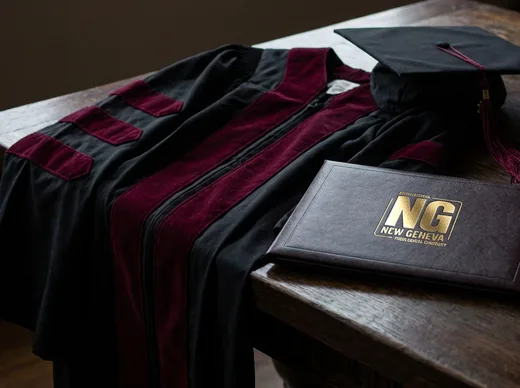 A New Geneva doctoral gown, mortarboard, and diploma folder resting on a wooden table.