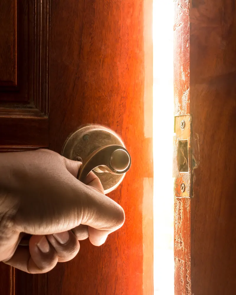A hand on a door handle, daylight showing through the opening door.