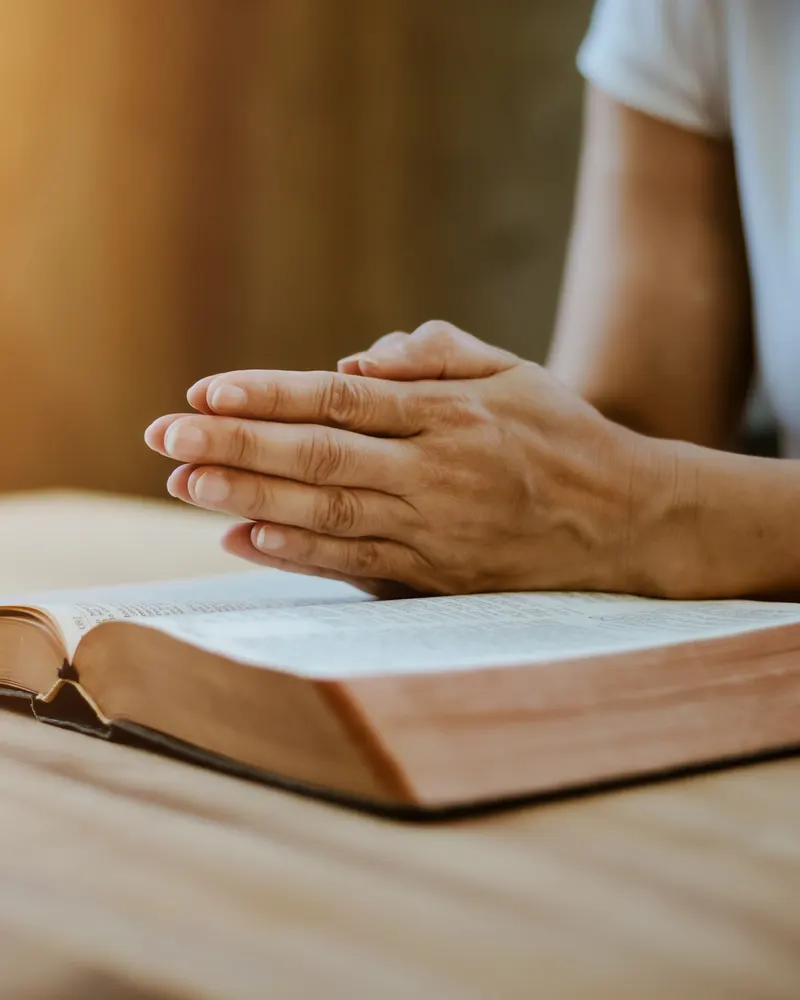 Hands folded in prayer resting on an open Bible on a wooden table.